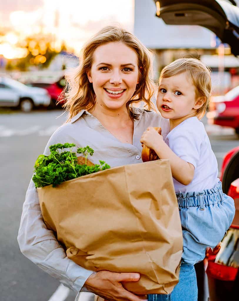 Smiling woman holds a toddler and a paper bag of fresh groceries in a sunlit parking lot, conveying warmth and everyday joy.