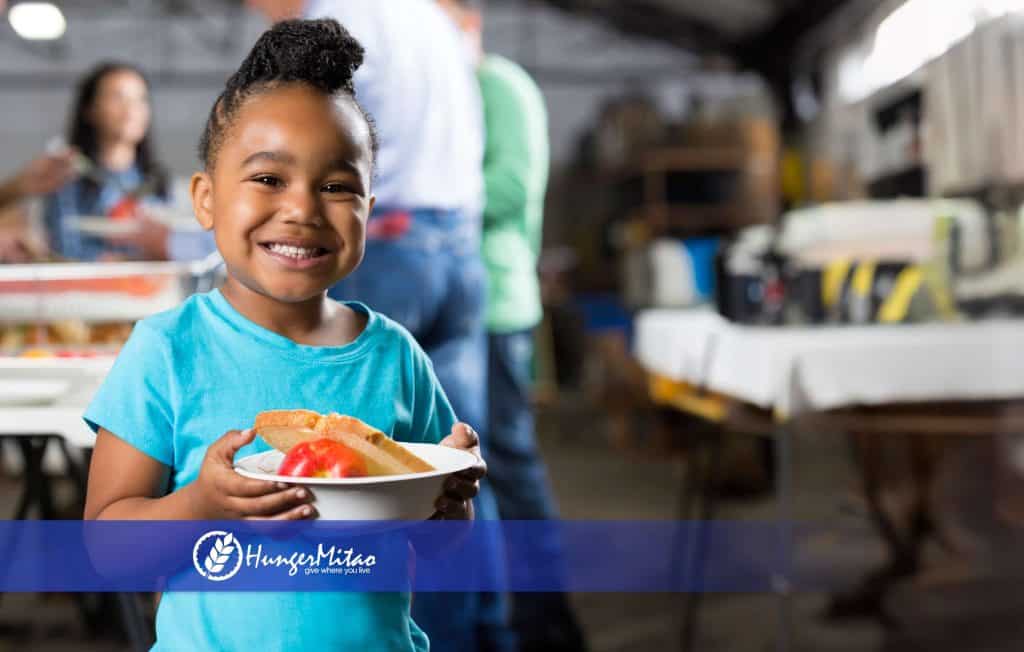 A smiling child in a turquoise shirt holds a plate with an apple and bread.